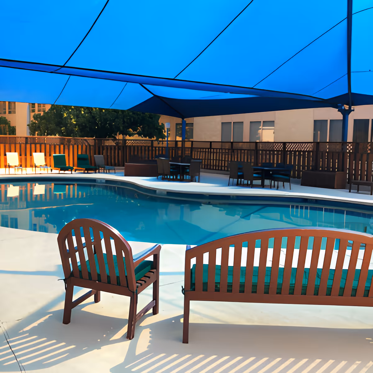 Outdoor swimming pool area with a blue shade canopy overhead, wooden chairs and benches with green cushions, and tables with chairs around the pool. A wooden fence and buildings are visible in the background.