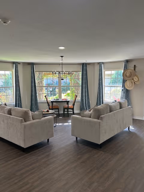 A bright living room area with two beige sofas facing each other on a wooden floor. Behind the sofas, there is a dining table with four chairs near large windows with blue curtains, allowing natural light to fill the room. A wall-mounted coat rack with hats is visible on the right side.