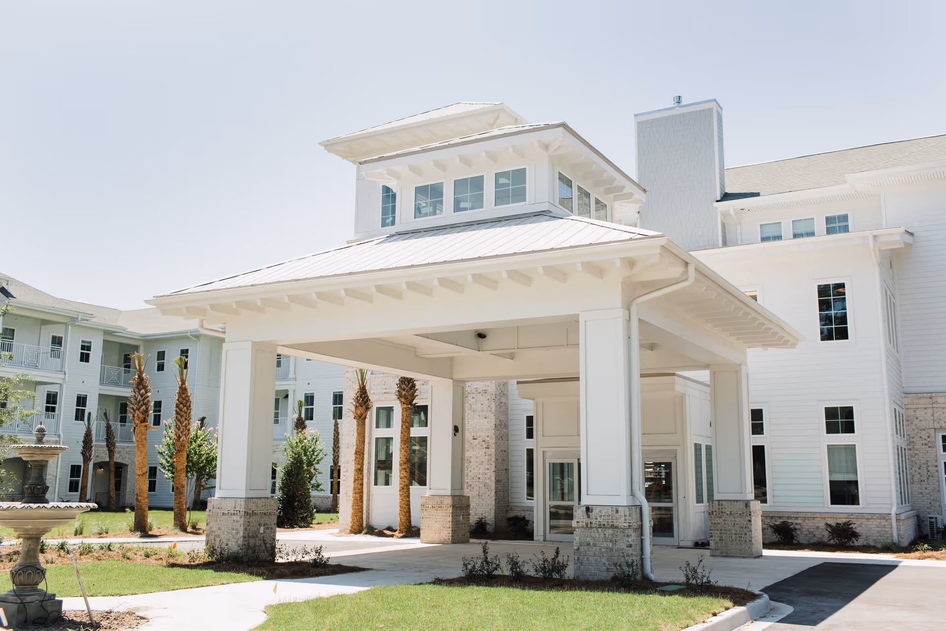 Exterior view of a large, white senior living facility building with a covered entrance supported by white columns. The building has multiple windows and a light-colored roof. There are palm trees and a fountain in the landscaped area near the entrance.