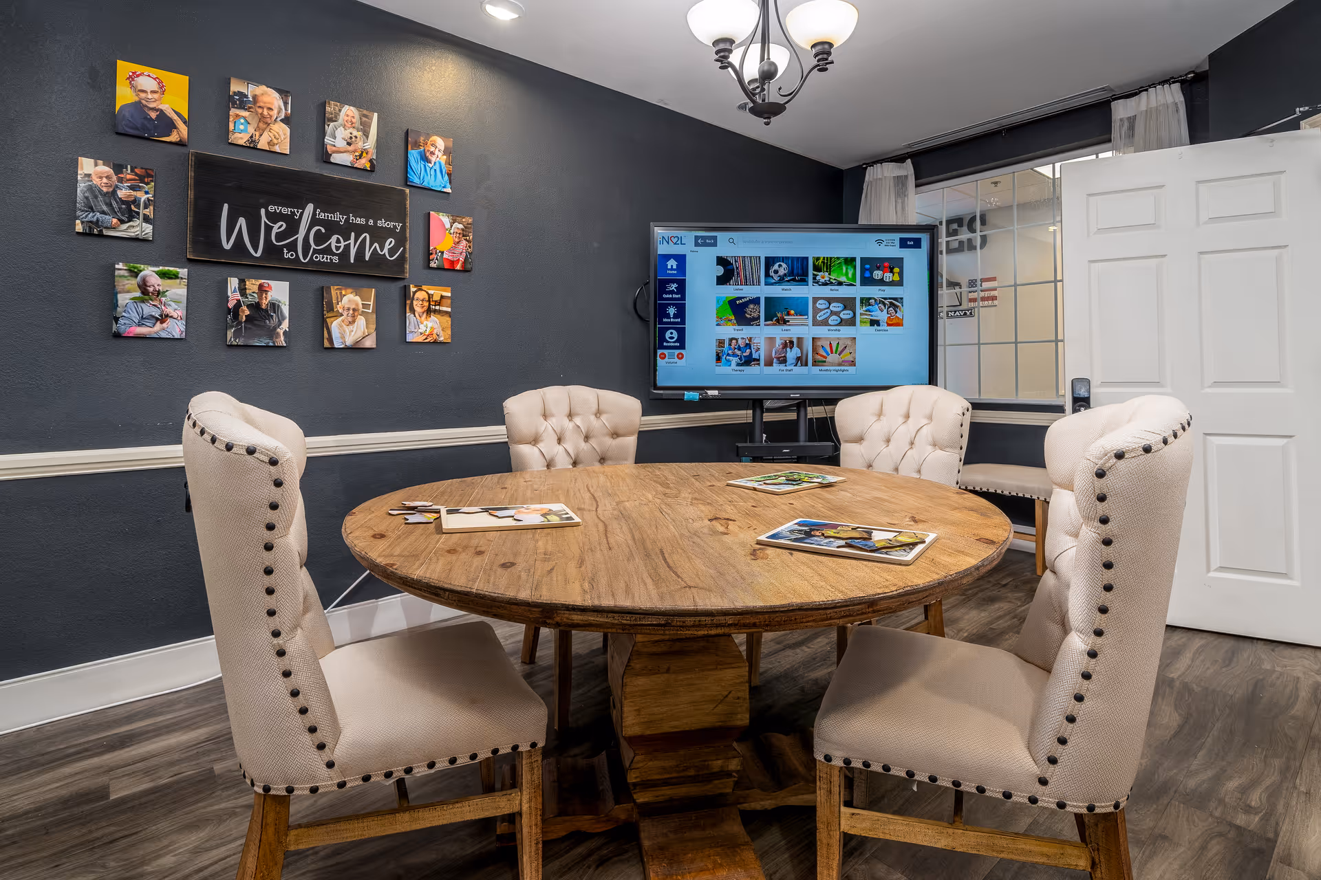 A cozy meeting or activity room with a round wooden table surrounded by five beige upholstered chairs with nailhead trim. On the dark gray wall behind the table, there is a collage of photos of elderly people surrounding a sign that reads 'every family has a story Welcome to ours.' A large flat-screen TV is mounted on a stand in the corner, displaying a colorful interface. The room has wood flooring, a white door, and a window with a view into another room.