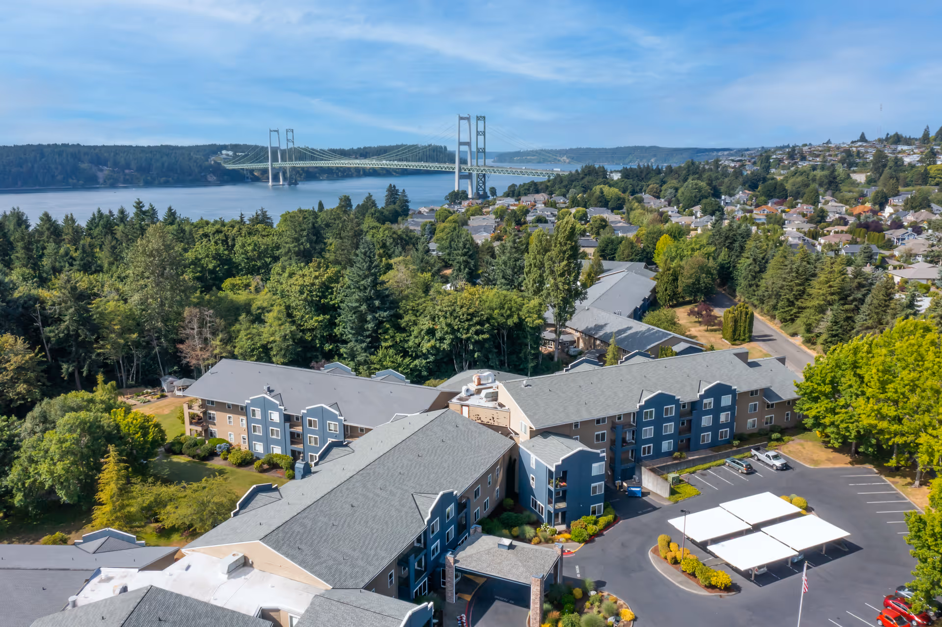Aerial view of Cogir at The Narrows senior living facility surrounded by trees and greenery, with a large suspension bridge and water body in the background under a partly cloudy sky.