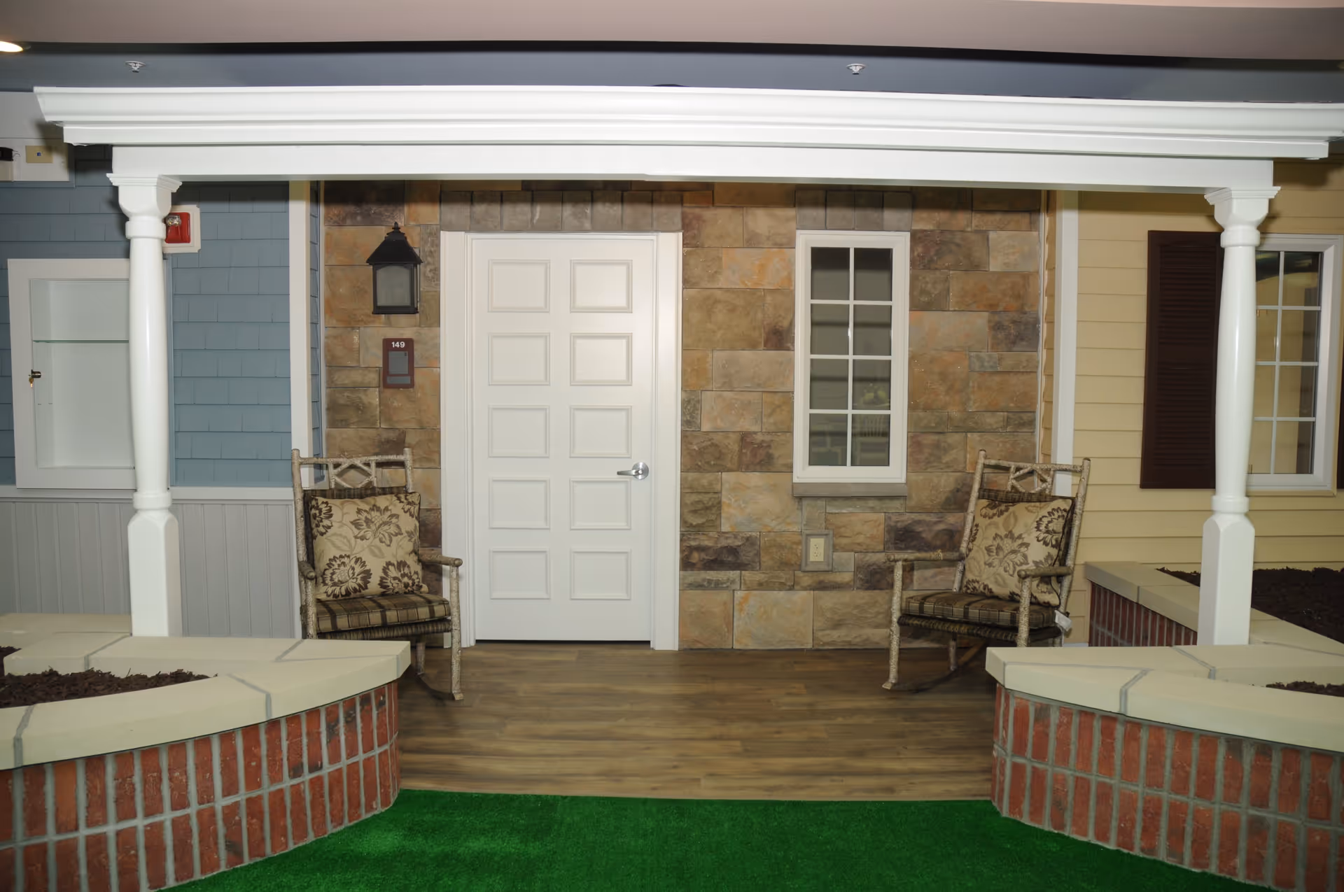 Covered porch area with a white door and a window on a stone wall. Two cushioned rocking chairs are placed on either side of the door. The porch has wooden flooring and is bordered by low brick planters with mulch. Green artificial grass is visible in the foreground.