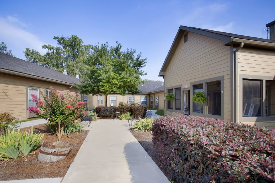 Outdoor courtyard area of a senior living facility with a concrete walkway, surrounded by bushes, flowering plants, and trees. Beige buildings with multiple windows and hanging plants are visible on both sides under a clear blue sky.
