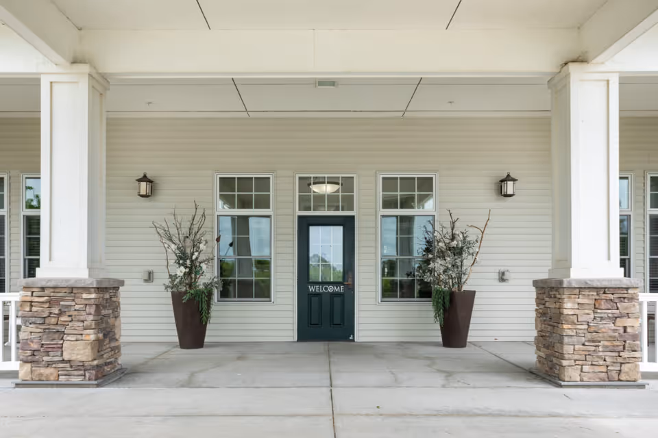 Front entrance of a building with a dark green door that has a 'WELCOME' sign on it, flanked by two tall windows and two large planters with decorative branches. The entrance is covered by a white ceiling supported by columns with stone bases.