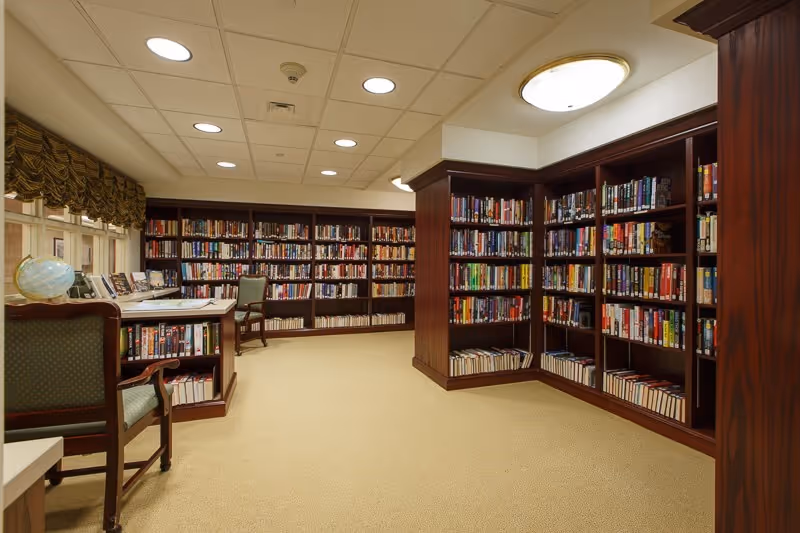 Interior of a library room with wooden bookshelves filled with books along the walls, a desk with a globe and chairs, and a beige carpeted floor under ceiling lights.