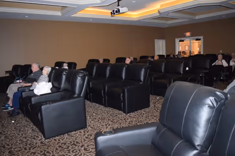 A room with multiple rows of black leather recliner chairs arranged in a theater-style seating. Several elderly people are seated, facing forward, suggesting they are watching a presentation or movie. The room has a patterned carpet, beige walls, and a ceiling-mounted projector.