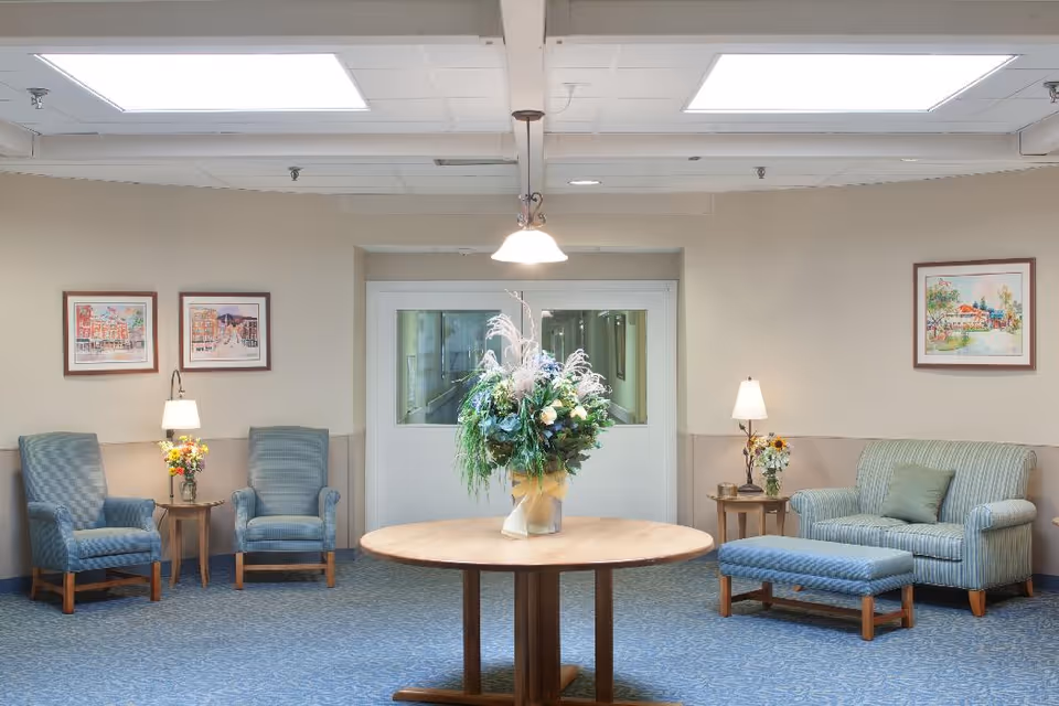 A sitting area in a senior living facility with two blue upholstered armchairs and a small wooden side table with a lamp and flowers on the left, a blue upholstered loveseat with a matching ottoman and a side table with a lamp and flowers on the right, and a round wooden table with a large floral arrangement in the center. The walls are beige with framed artwork, and there are double glass doors in the background.