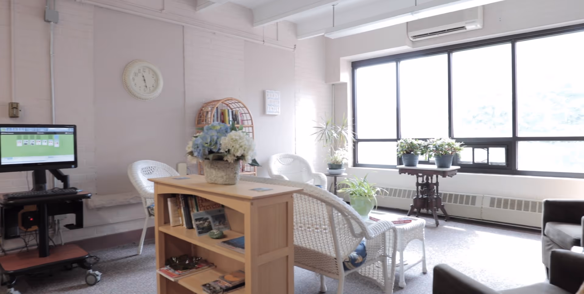 A bright and cozy living room area with large windows letting in natural light. The room features white wicker chairs, a small wooden bookshelf with books and magazines, several potted plants, a wall clock, and a computer on a mobile stand displaying a solitaire game.