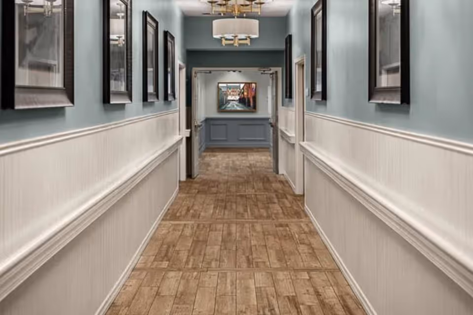 A long, well-lit hallway with light blue walls and white wainscoting. The floor is covered with wood-patterned tiles. Several framed pictures hang on both sides of the hallway. At the end of the hallway, there is an open doorway leading to another room with a painting visible on the far wall. A modern ceiling light fixture is mounted above.