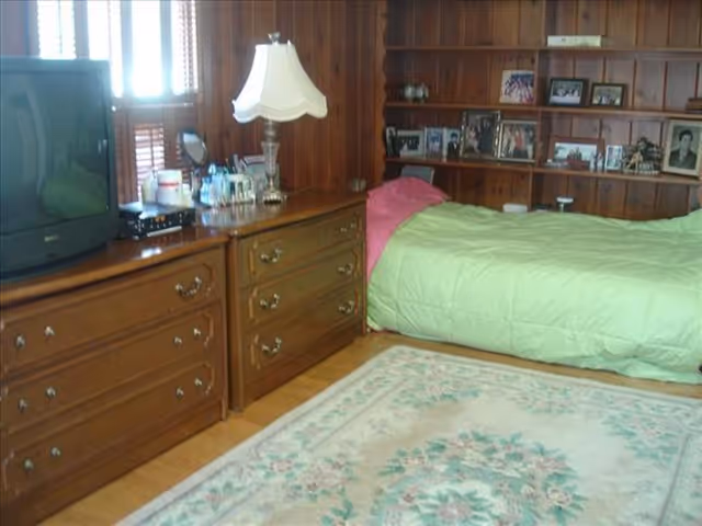 A cozy bedroom with wood-paneled walls, a bed with a green comforter and pink pillow, a wooden dresser with a lamp and various items on top, and a TV. There are shelves above the bed displaying framed photos and decorative items. A floral area rug covers part of the wooden floor.