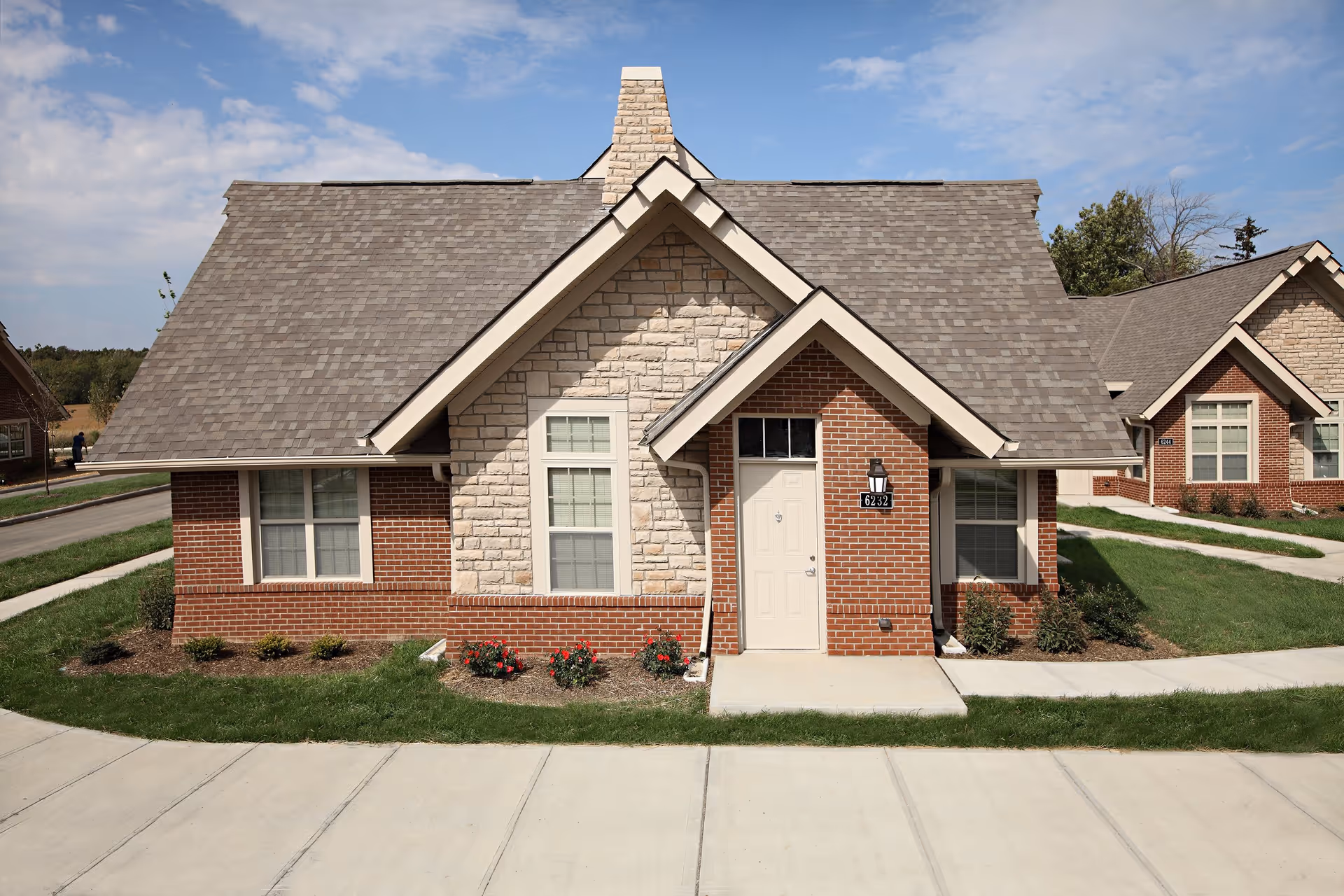 Front exterior view of a single-story brick and stone residential building with a gray shingled roof, a white door, and windows on either side. There are small flower beds with red flowers and shrubs in front of the building, and a concrete sidewalk and driveway in the foreground under a partly cloudy sky.