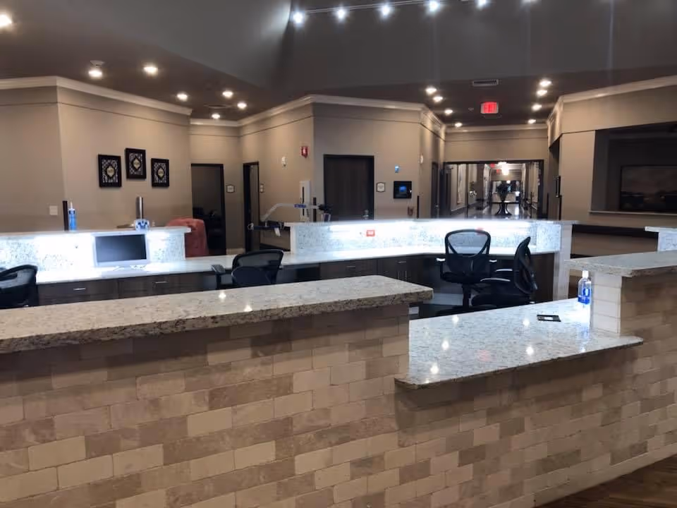 Interior view of a senior living facility reception or nurse station area with a long granite countertop, office chairs, computer monitors, and a hallway leading to other rooms. The walls are beige with framed artwork and recessed lighting on the ceiling.