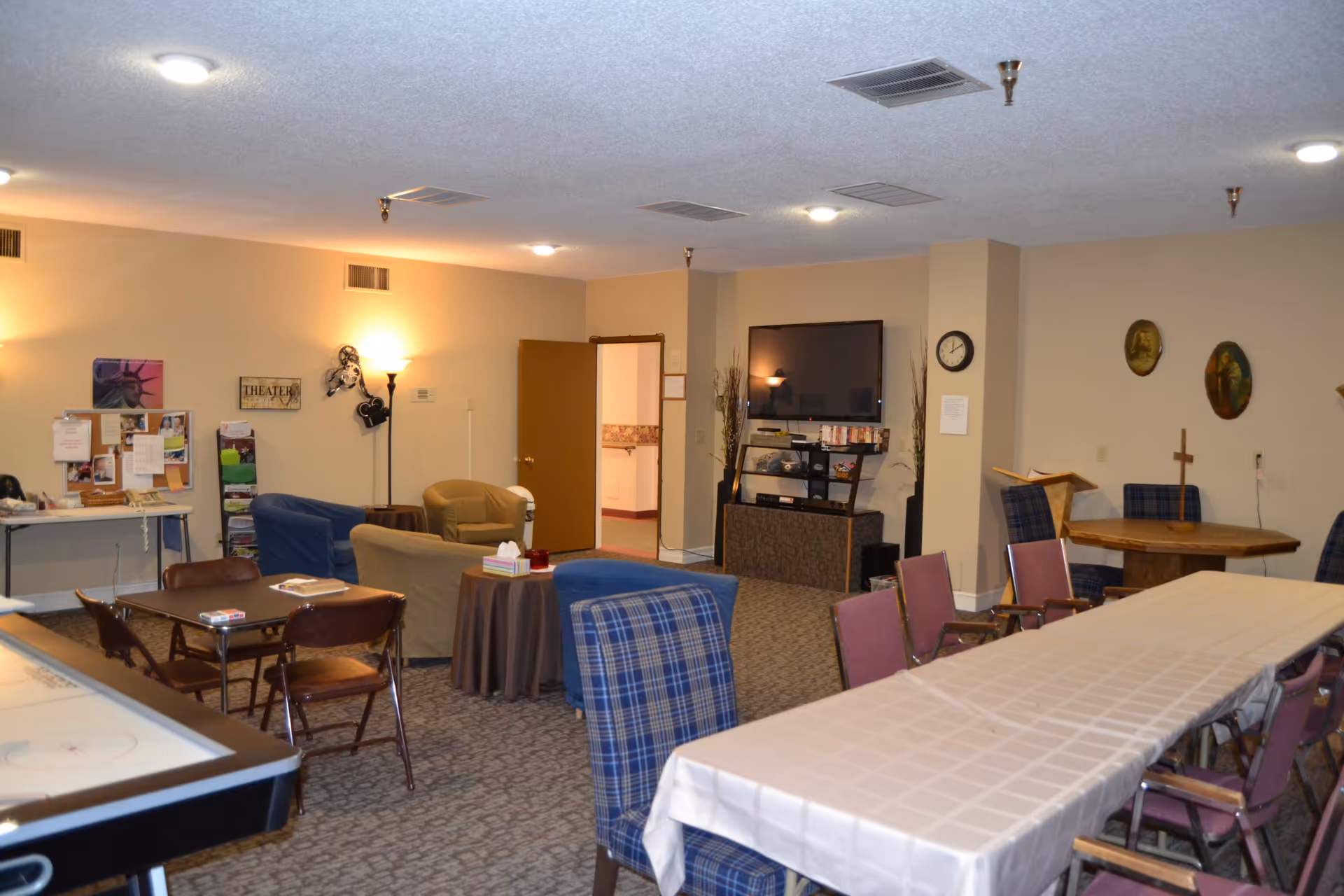 A common area in a senior living facility with multiple seating arrangements including armchairs, tables with chairs, and a long table covered with a white tablecloth. The room has beige walls, carpeted floor, a flat-screen TV mounted on the wall, and various decorations including wall art and a bulletin board.
