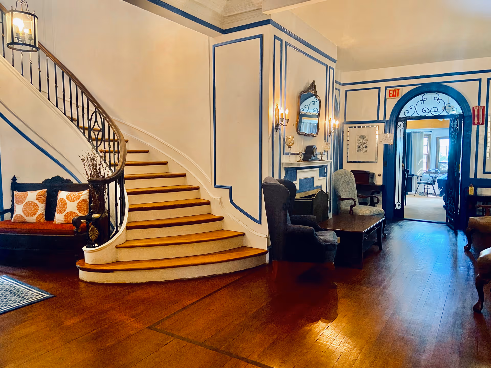 A bright, elegant interior lobby with a curved wooden staircase, upholstered chairs, a small fireplace, and an arched doorway.