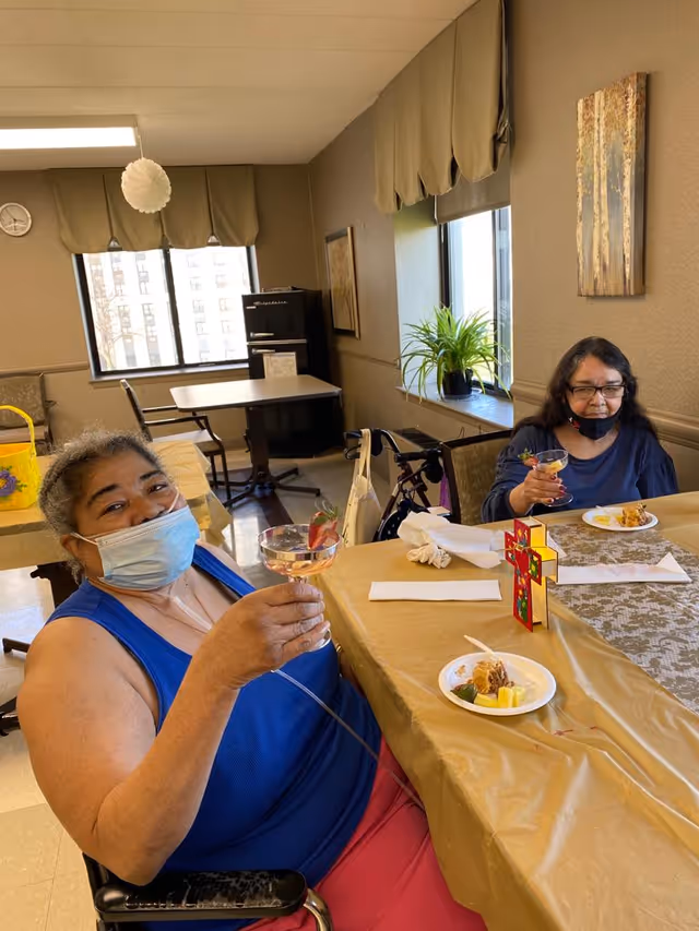 Two elderly women sitting at a table in a dining area, each holding a glass with a strawberry garnish. One woman is wearing a blue sleeveless top and a face mask, while the other is wearing a dark top with her mask pulled down. There are plates with food on the table, a decorative cross, a plant on the windowsill, and a small refrigerator in the background.