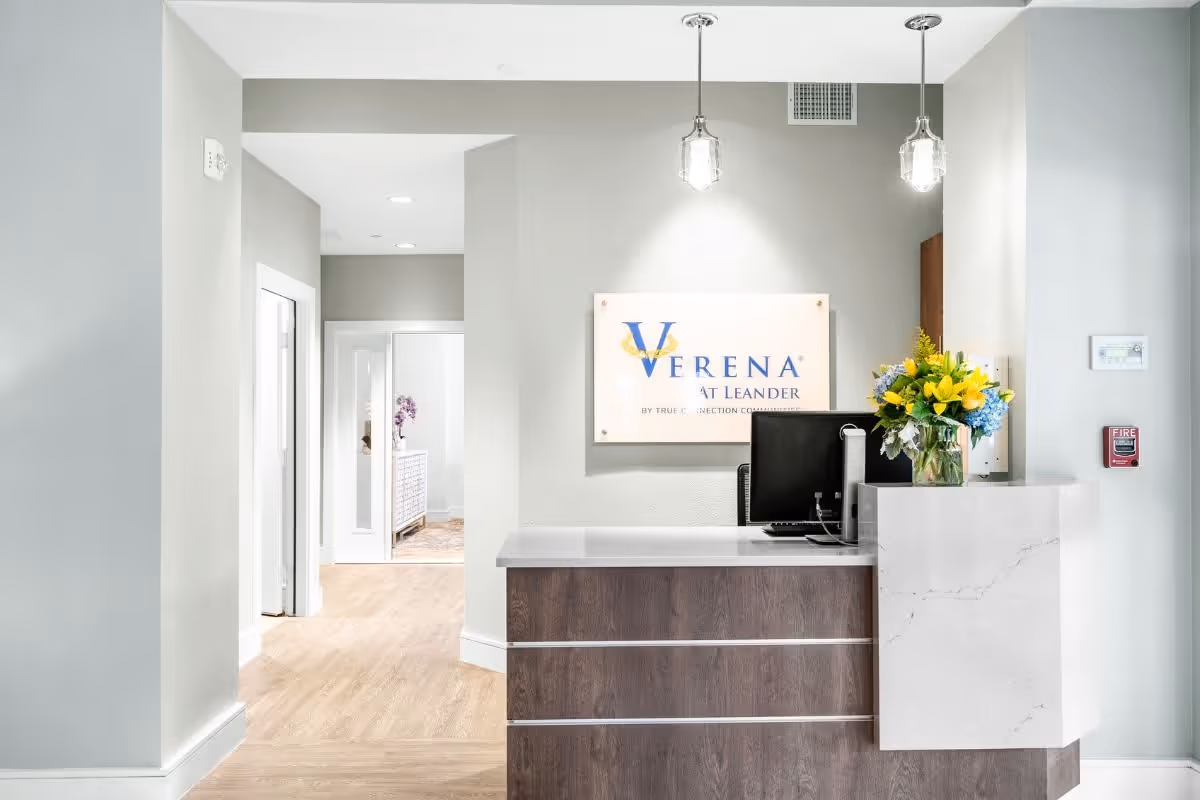 Reception area of Verena at Leander facility with a modern desk, computer monitor, and a vase of yellow and blue flowers. The wall behind the desk features a sign with the facility's name. The space has light-colored walls and wood flooring, with a hallway leading to other rooms.