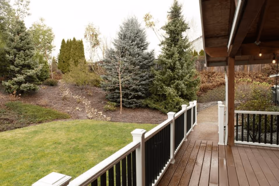 View of a backyard garden area with green grass, various evergreen trees, and a wooden deck with white and black railing. A covered porch with hanging lights is visible on the right side, along with a concrete pathway leading into the garden.