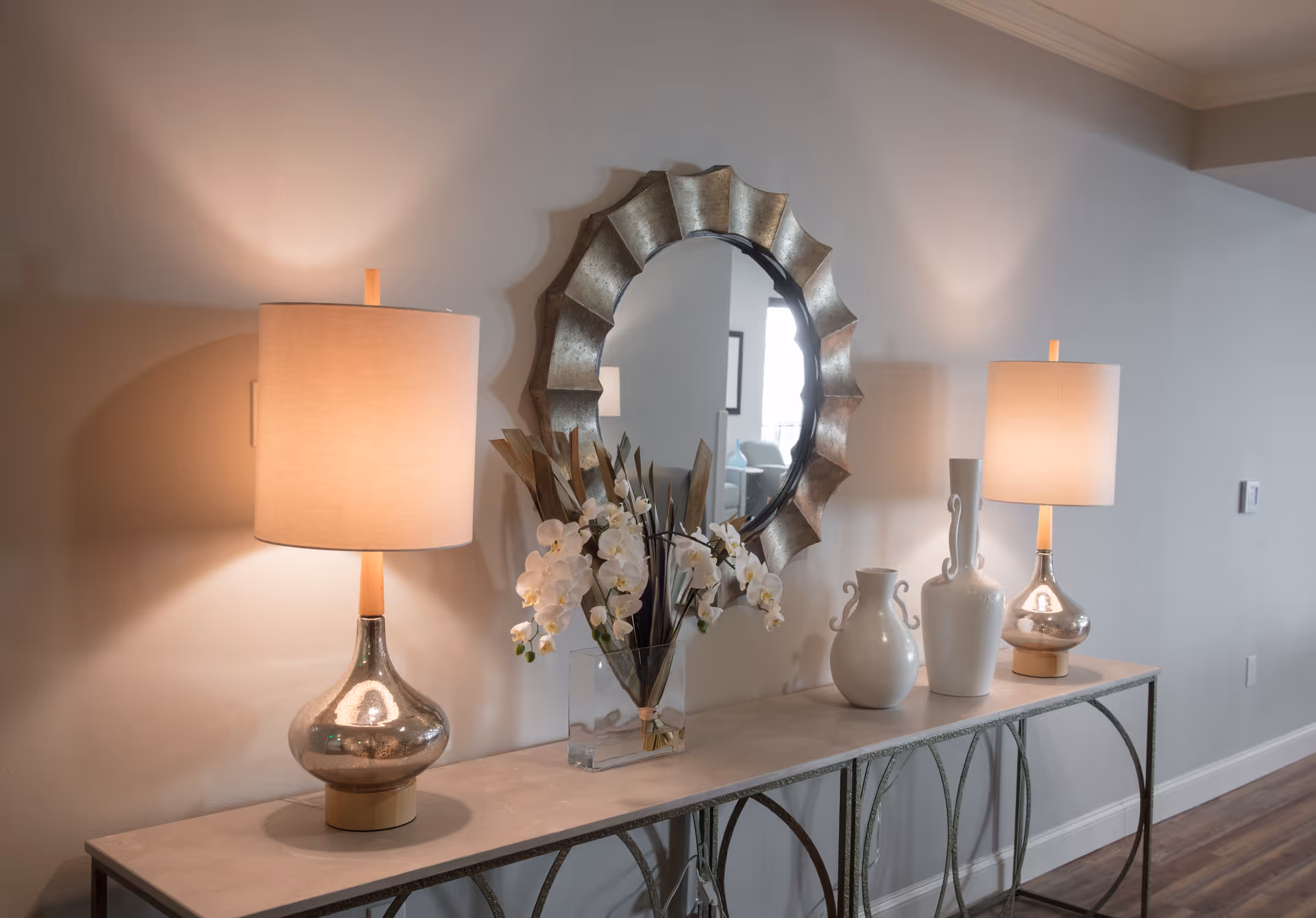 A decorative console table with two lit table lamps, white ceramic vases, and a glass vase with white orchids and dried leaves, positioned against a light-colored wall with a round, scalloped-edge mirror hanging above it.