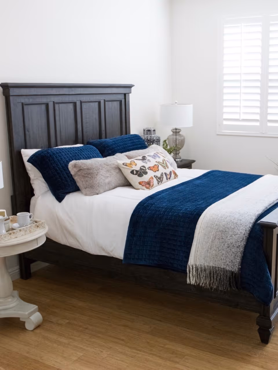 A neatly made bed with a dark wooden headboard, white sheets, blue quilted blanket, and decorative pillows including one with butterfly designs. Next to the bed is a small round side table with a tray holding a teapot and cups. A bedside table with a lamp and a small plant is visible near a window with white shutters.