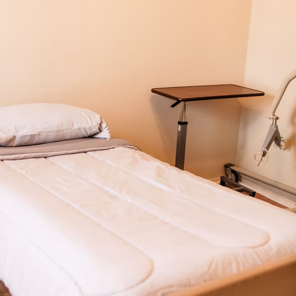 A neatly made single bed with a white pillow and a light gray blanket in a simple room. Next to the bed is an adjustable overbed table and a bed assist rail attached to the wall.