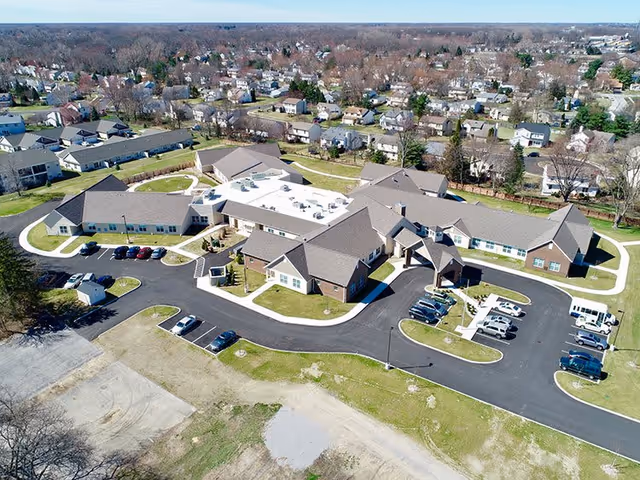 Aerial view of Charter Senior Living of Oak Openings facility showing multiple connected buildings with pitched roofs, surrounding parking lots with cars, and landscaped green areas. Residential neighborhood with houses and trees is visible in the background.