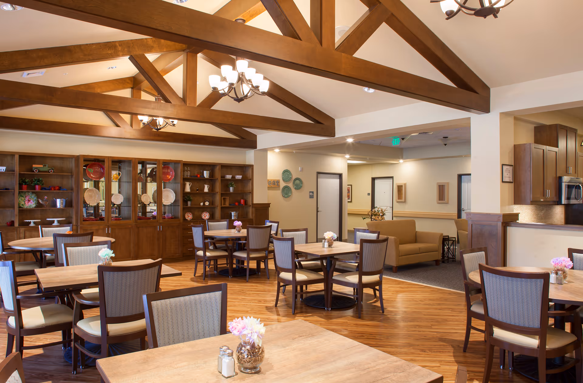 A spacious dining room in a senior living facility with wooden tables and cushioned chairs arranged neatly. The room features wooden ceiling beams, a chandelier, and a wooden cabinet displaying decorative plates and items. In the background, there is a small seating area with a couch and a view into a kitchen area with wooden cabinets and stainless steel appliances.