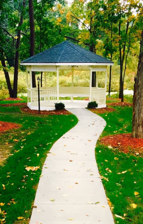 A white gazebo with a dark shingled roof at the end of a curved concrete pathway surrounded by green grass, trees, and landscaped mulch areas in an outdoor garden setting.