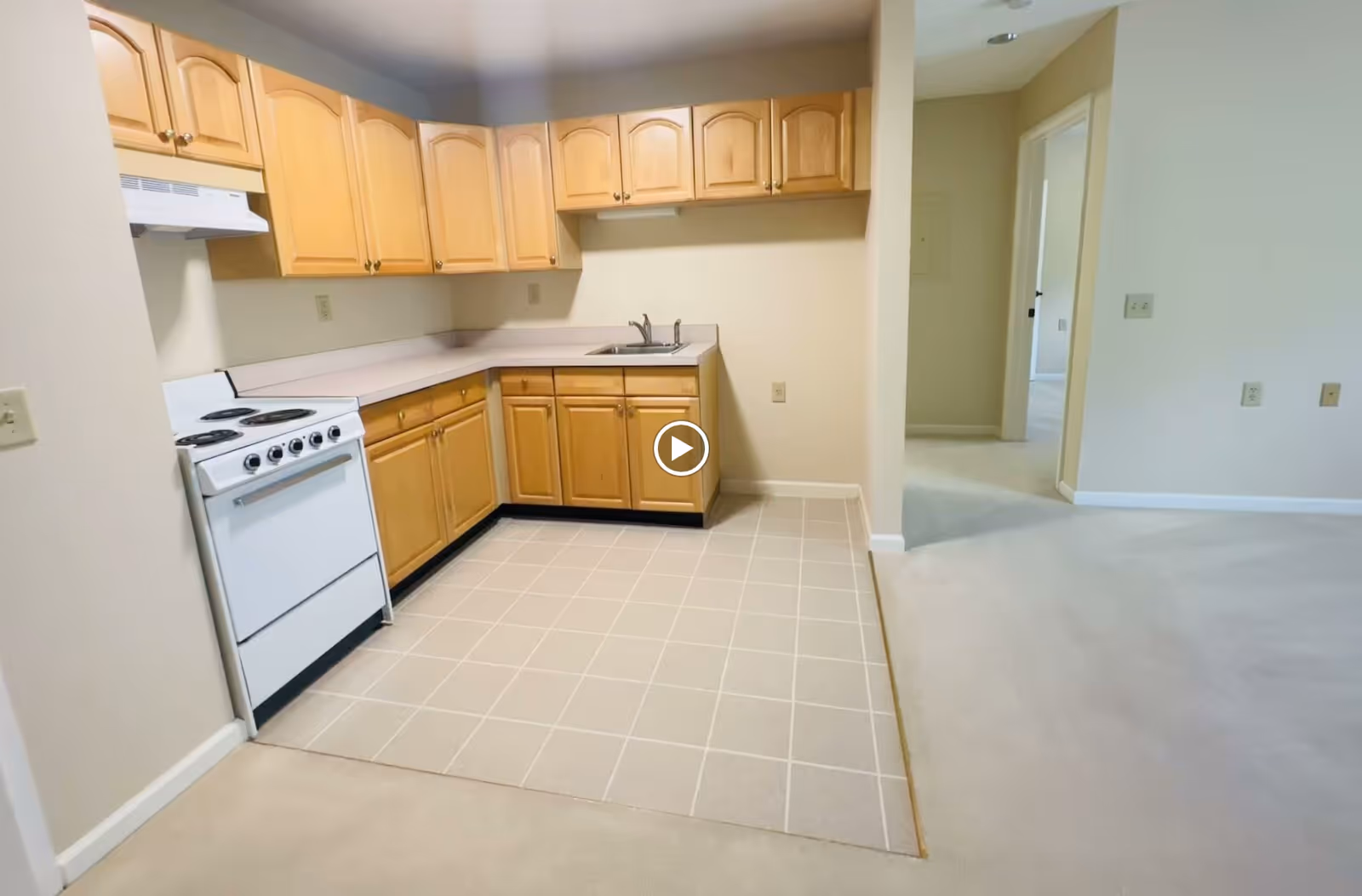 Small kitchen with light wood cabinets, a white electric stove and sink over a tiled floor opening into a carpeted living area.
