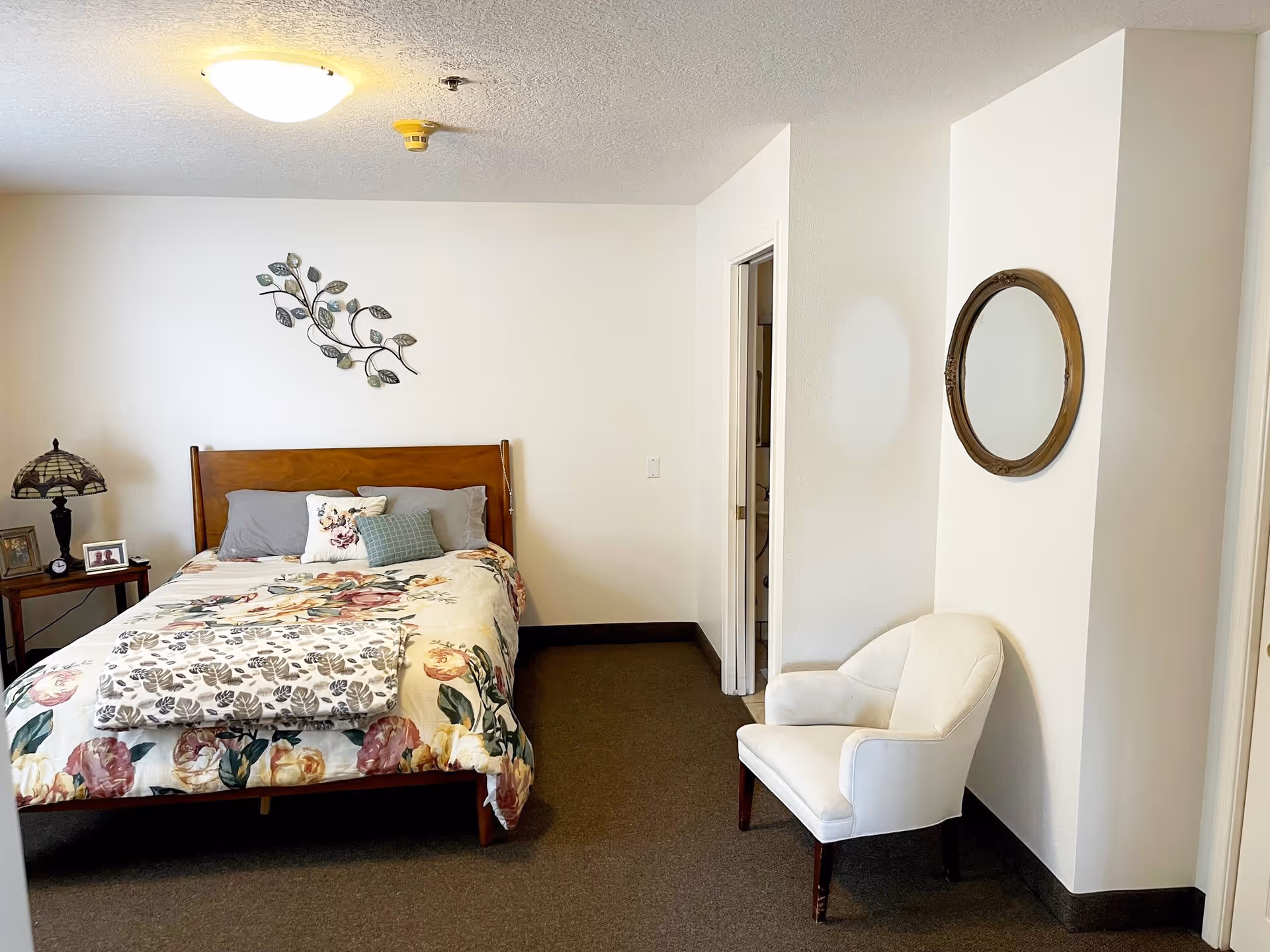 A cozy bedroom with a wooden bed frame, floral bedspread, and several pillows. To the left of the bed is a small wooden nightstand with a decorative lamp and framed photos. On the right side of the room, there is a white upholstered armchair beneath a round wall mirror. The walls are white, and the carpet is dark brown. A doorway leads to another room or bathroom.