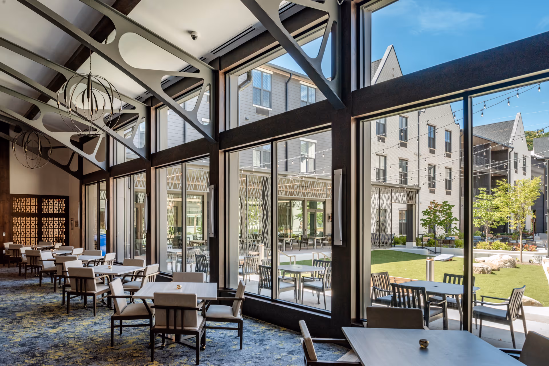 Interior dining area with multiple tables and chairs arranged neatly, large floor-to-ceiling windows providing a view of an outdoor courtyard with additional seating and greenery under a clear blue sky.
