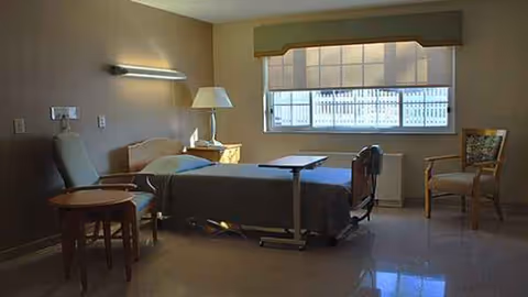 Patient bedroom in a care center with bed, chairs, and window.