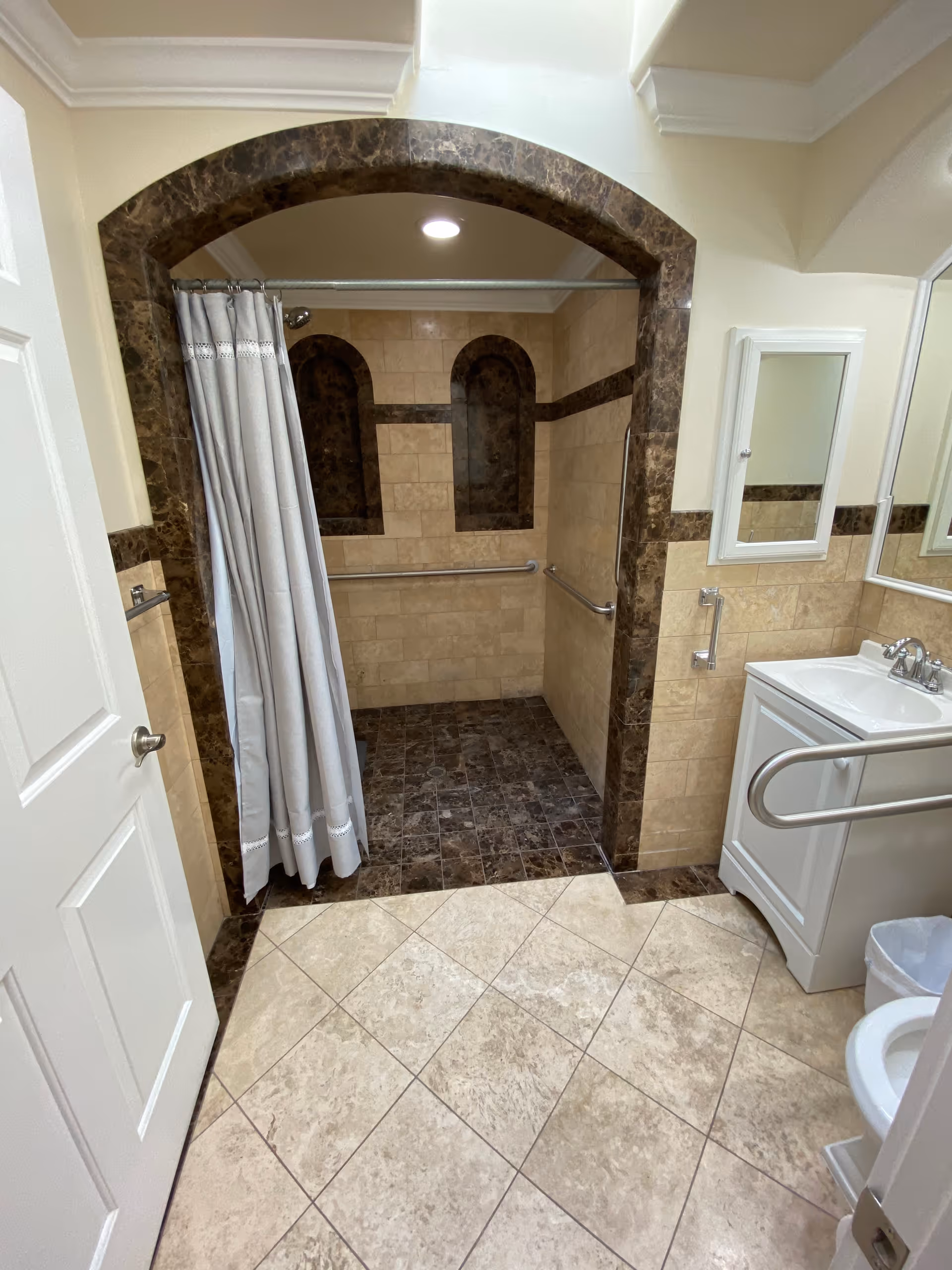 A bathroom with beige and brown tiled floors and walls. There is a walk-in shower area with a curved archway and a gray shower curtain. The shower area has grab bars installed on the walls. To the right, there is a white sink with a mirror above it and a grab bar next to the sink. A white door is partially open on the left side of the image.