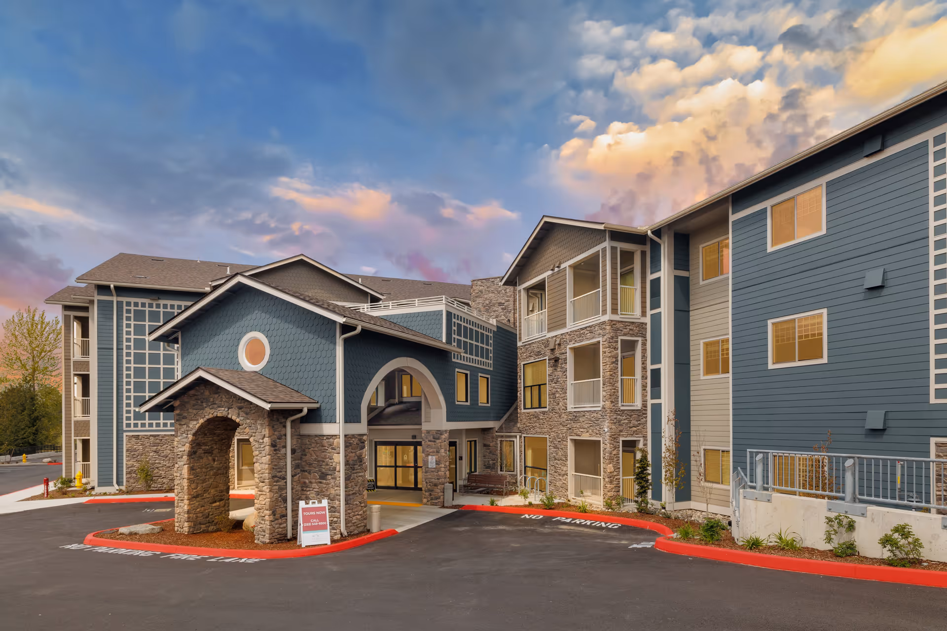 Exterior view of a senior living facility building named Cogir of Kent during sunset, featuring a stone and blue siding facade with multiple windows and a covered entrance with stone pillars. The sky is partly cloudy with warm colors.