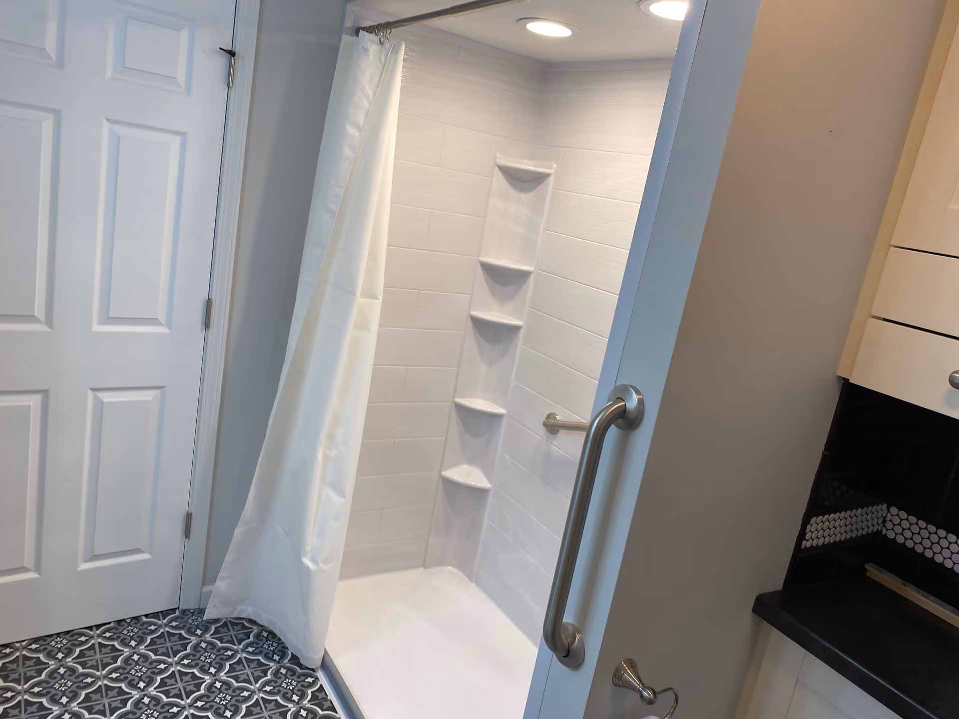 A clean, modern shower area with white tiled walls and built-in corner shelves. The shower has a white curtain partially drawn to the side and a sturdy metal grab bar on the wall outside the shower. The floor outside the shower features patterned tiles in shades of gray and white. A white door is visible to the left, and part of a kitchen or cabinet area is visible to the right.