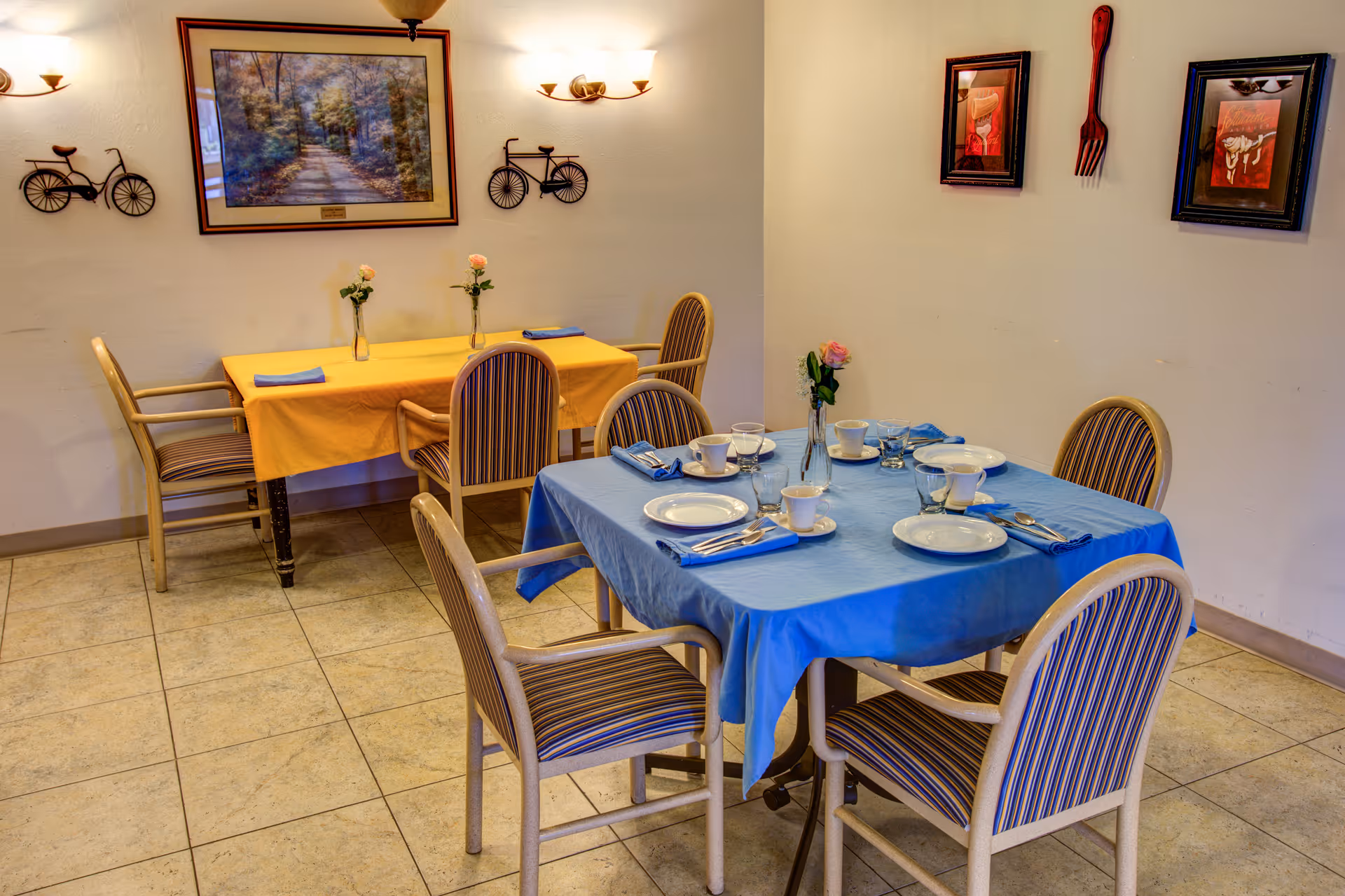 A small dining area with two tables covered in yellow and blue tablecloths. Each table is set with plates, cups, glasses, and silverware, and has a vase with a single pink rose. The chairs around the tables have striped upholstery. The walls are decorated with framed pictures and bicycle-shaped wall art.
