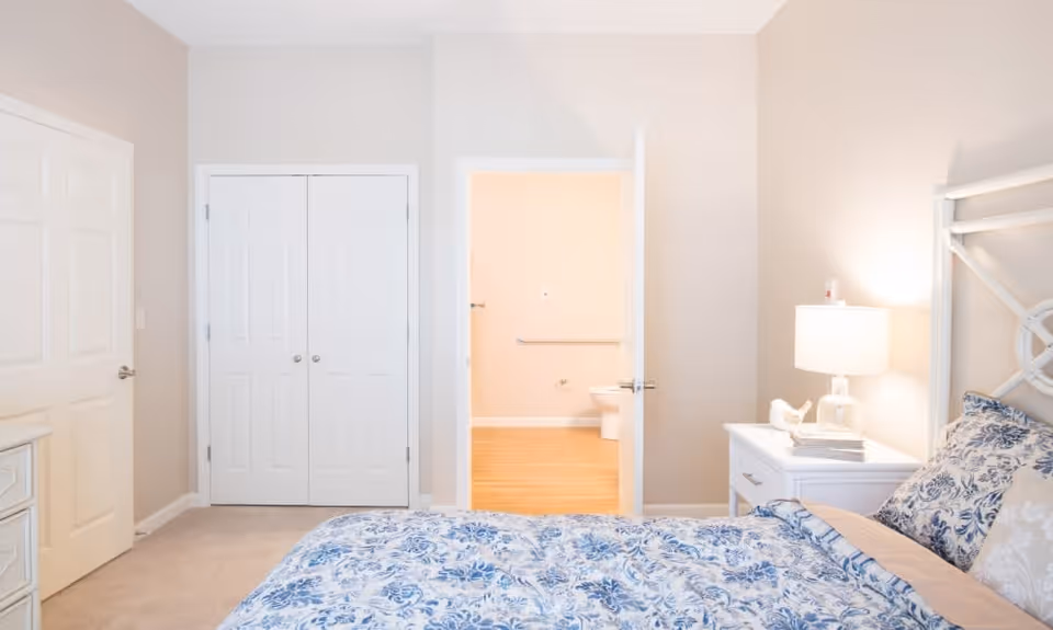 A bedroom with a bed covered in blue and white floral bedding, a white nightstand with a lamp and books, beige walls, and an open door leading to a bathroom with a visible toilet and grab bar.