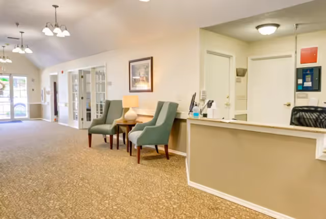 Bright reception lobby with a front desk, two upholstered chairs and a lamp next to an entrance.