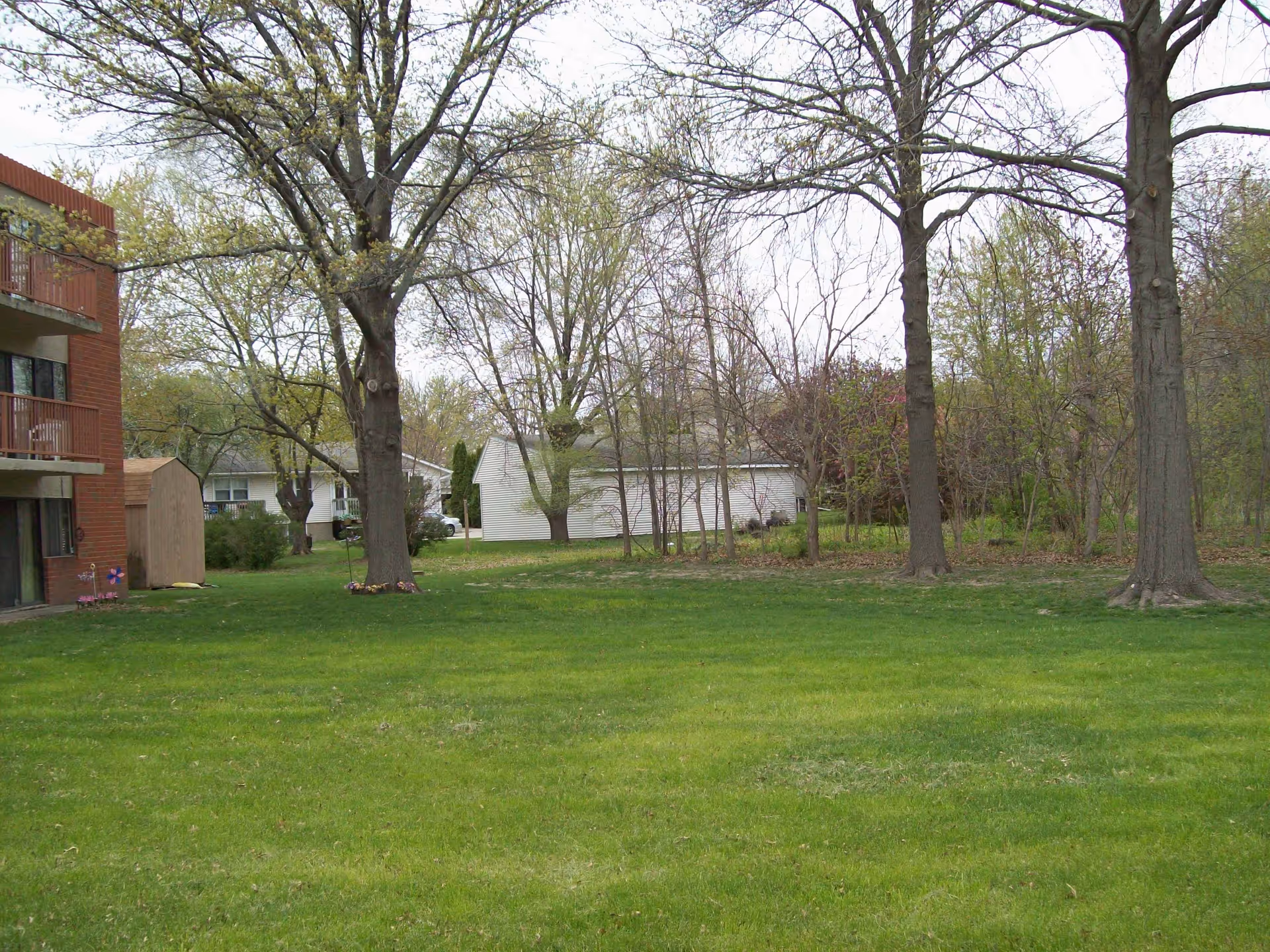 A grassy outdoor area with several large trees and a few small flower beds near the base of the trees. On the left side, there is a multi-story brick building with balconies and a small shed. In the background, there are residential houses and more trees with sparse foliage.