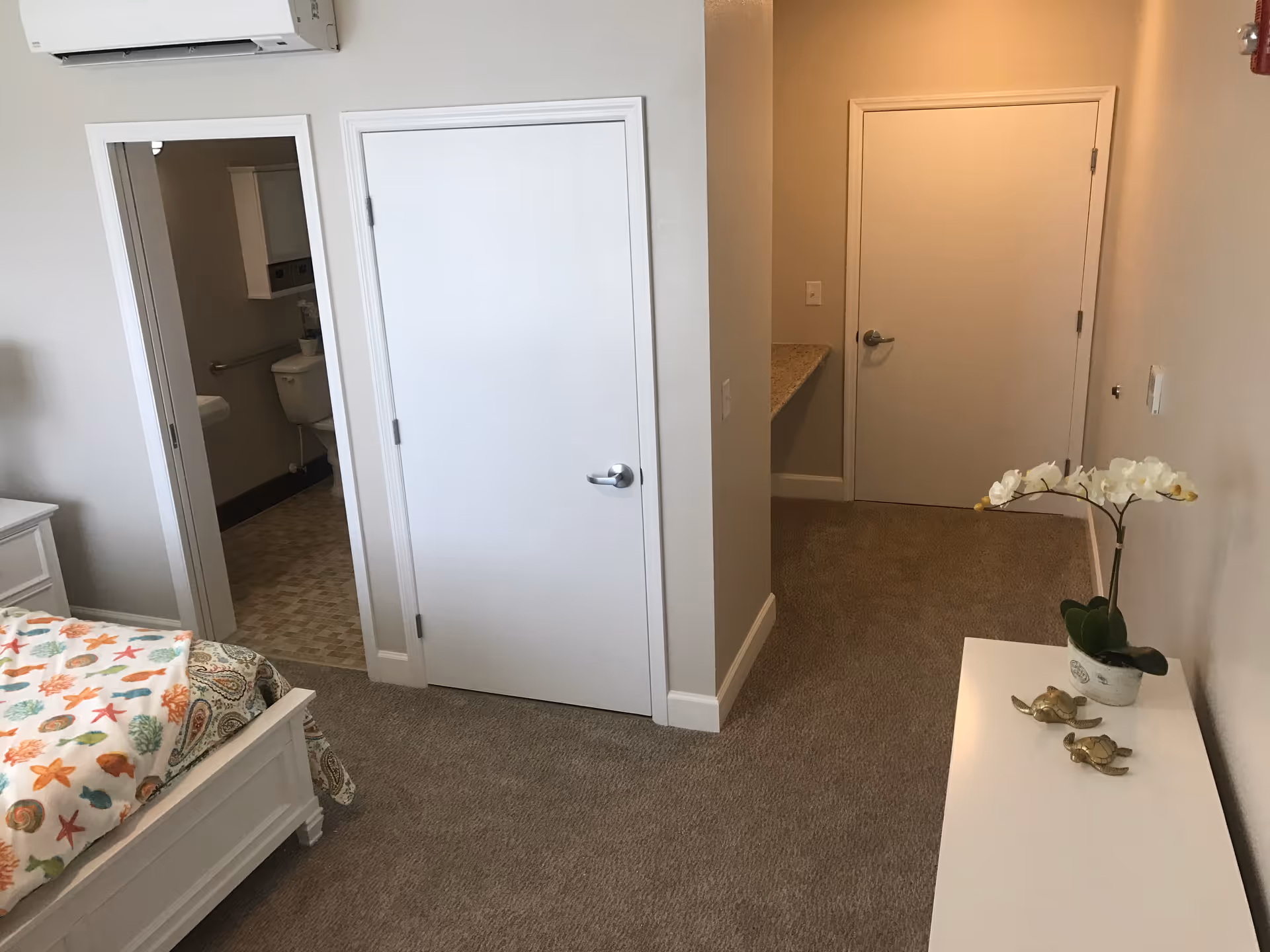 A bedroom with a white bed frame and colorful seashell-patterned bedding. There is a white dresser with a potted orchid and two small turtle figurines on top. The room has beige carpet and light-colored walls. Two white doors are visible, one leading to a bathroom with a toilet and sink, and the other likely a closet or another room. An air conditioning unit is mounted on the wall above the bathroom door.