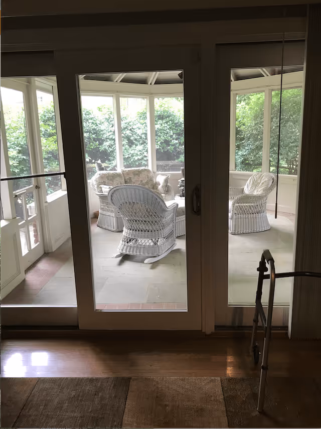 View through glass doors into a sunroom or enclosed porch with white wicker furniture including a rocking chair, loveseat, and armchair with floral cushions. Outside greenery is visible through the windows. A walker is partially visible inside near the doors.