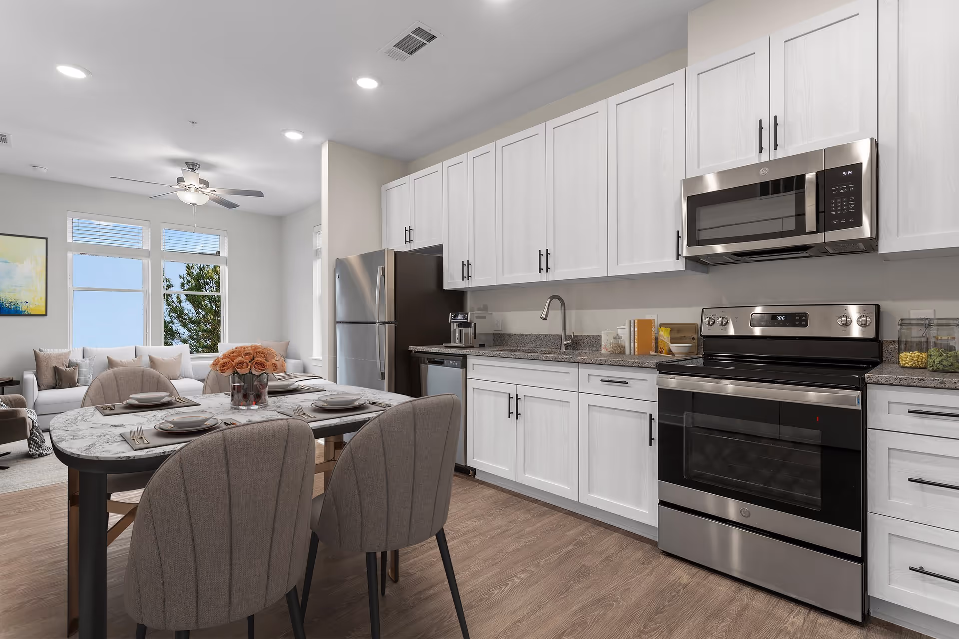 Modern kitchen and dining area with white cabinets, stainless steel appliances including a refrigerator, dishwasher, stove, and microwave. A dining table with four upholstered chairs is set with plates and cutlery, and a vase of pink roses is in the center. In the background, a living area with a white sofa, pillows, a ceiling fan, and large windows letting in natural light is visible.
