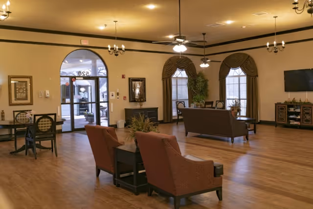 A spacious senior living facility common area with wooden floors, multiple seating arrangements including armchairs and sofas, large windows with draped curtains, ceiling fans with lights, chandeliers, and a wall-mounted TV. The entrance door with glass panels is visible in the background.
