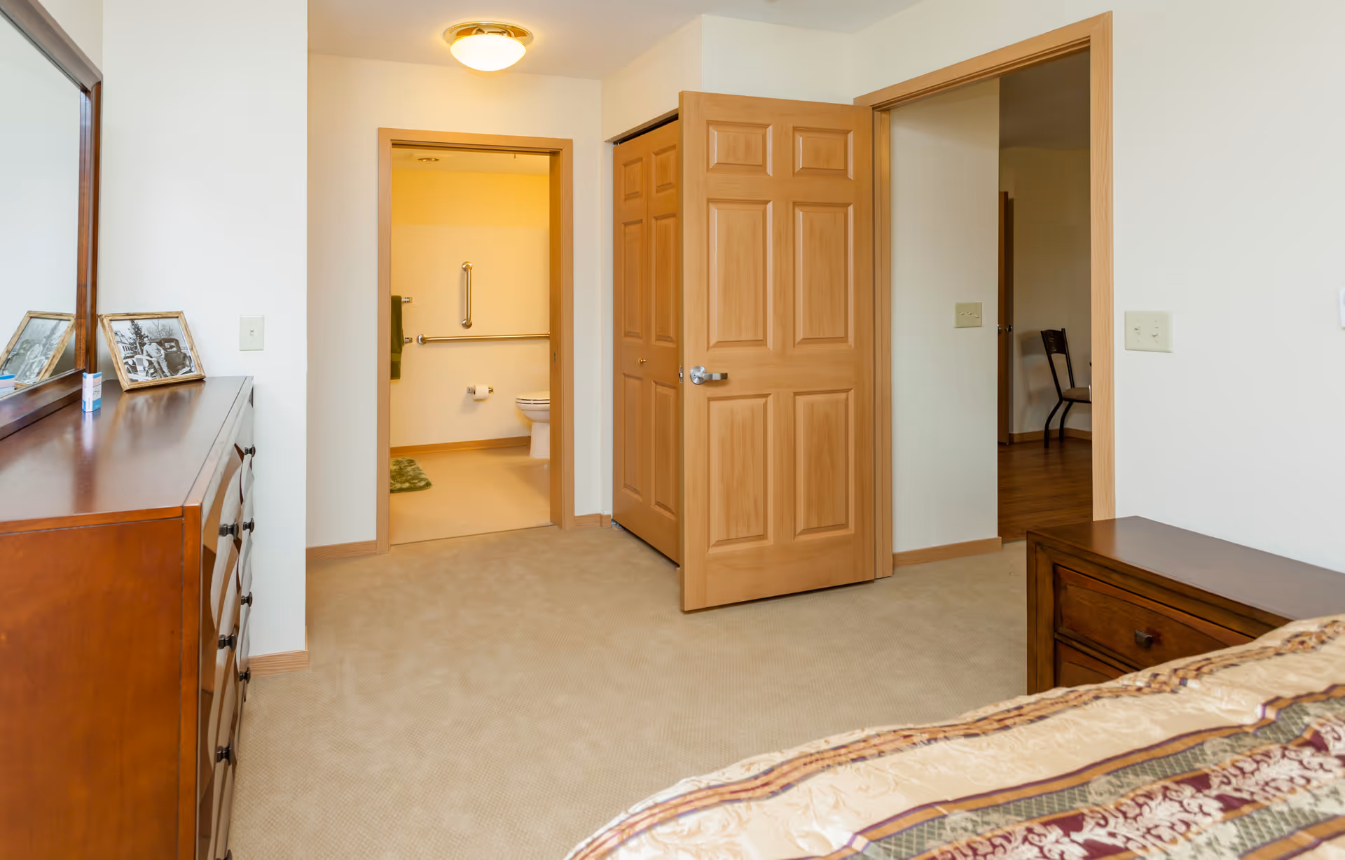 View of a bedroom with beige carpet, wooden dresser with a mirror and framed photos on top, a partially visible bed with patterned bedding, and two open doors leading to a bathroom with a toilet and grab bars, and another room with a wooden chair.