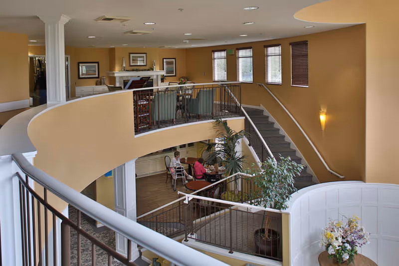 Multi-level, warmly lit interior common area with curved staircases, railings, seating tables, plants, and two residents seated at a table.