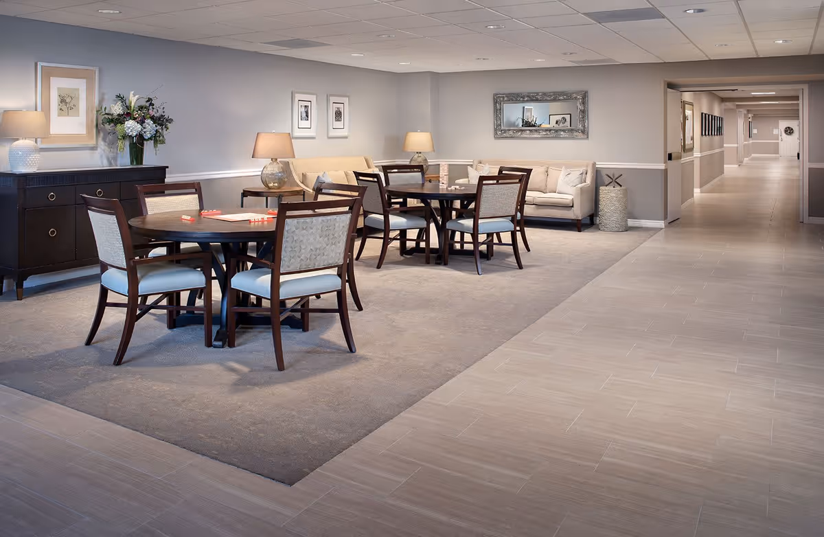 A senior living facility common area with two round tables surrounded by wooden chairs with cushioned seats. There are two beige sofas along the wall, two table lamps on side tables, framed artwork on the walls, and a decorative flower arrangement on a dark wooden sideboard. The floor is tiled with a carpeted area under the tables and sofas. A long hallway extends to the right.