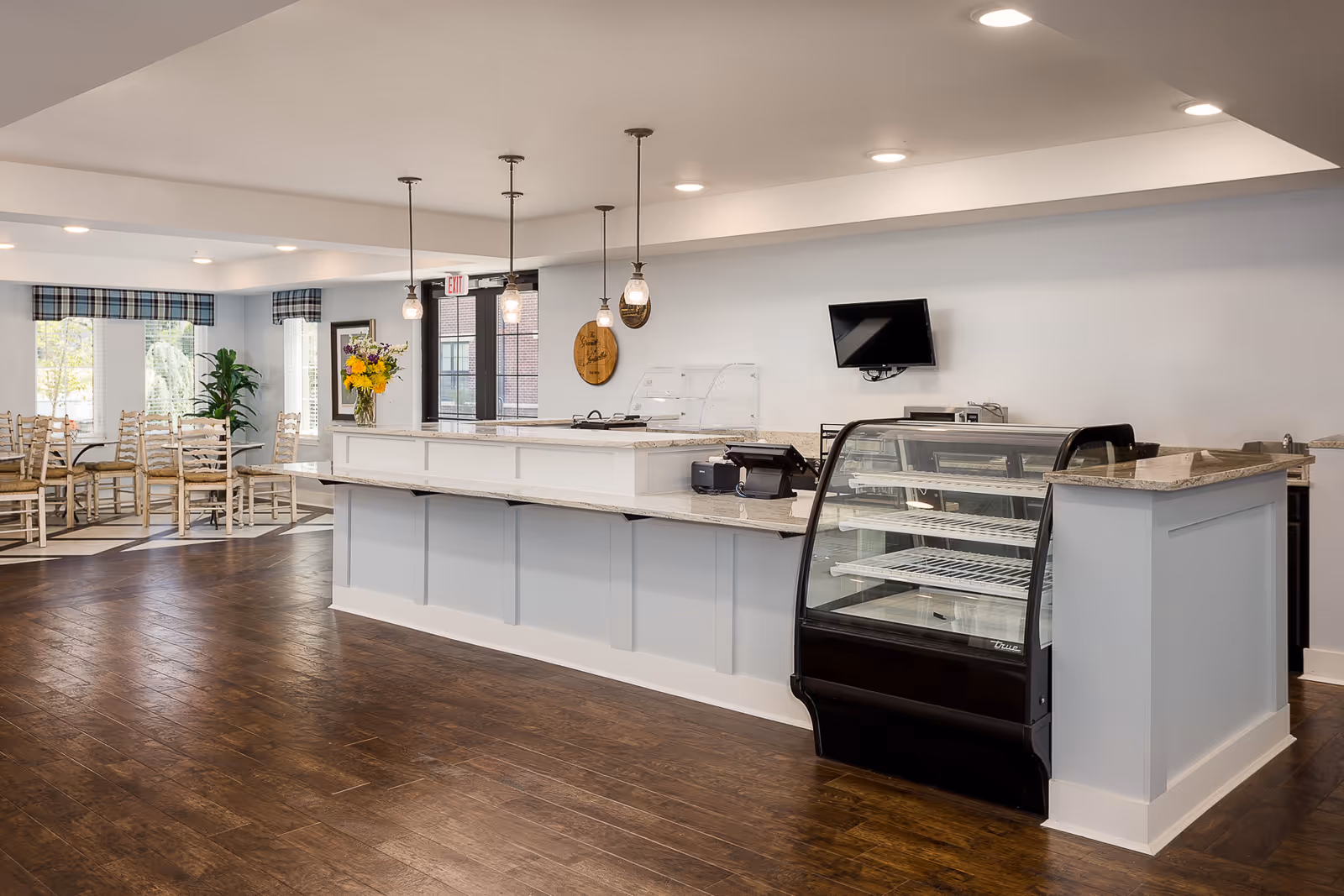 Interior view of a senior living facility dining area with a long counter featuring a glass display case, pendant lights hanging above, and wooden chairs and tables near large windows with plaid valances. A small TV is mounted on the wall behind the counter.