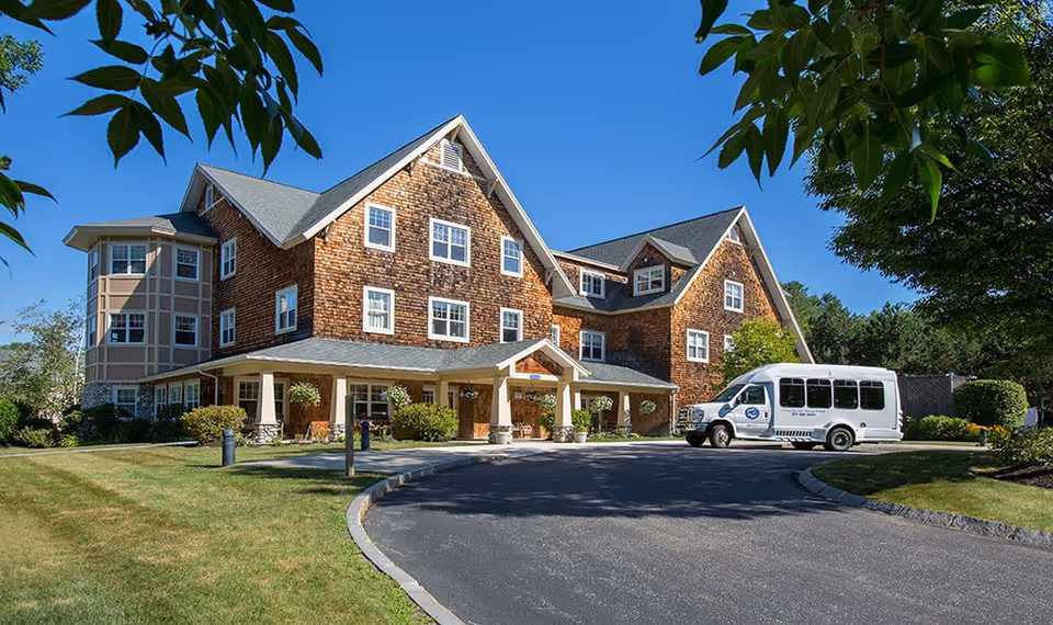 Brick senior living facility with a curved driveway, entrance, and white shuttle bus.
