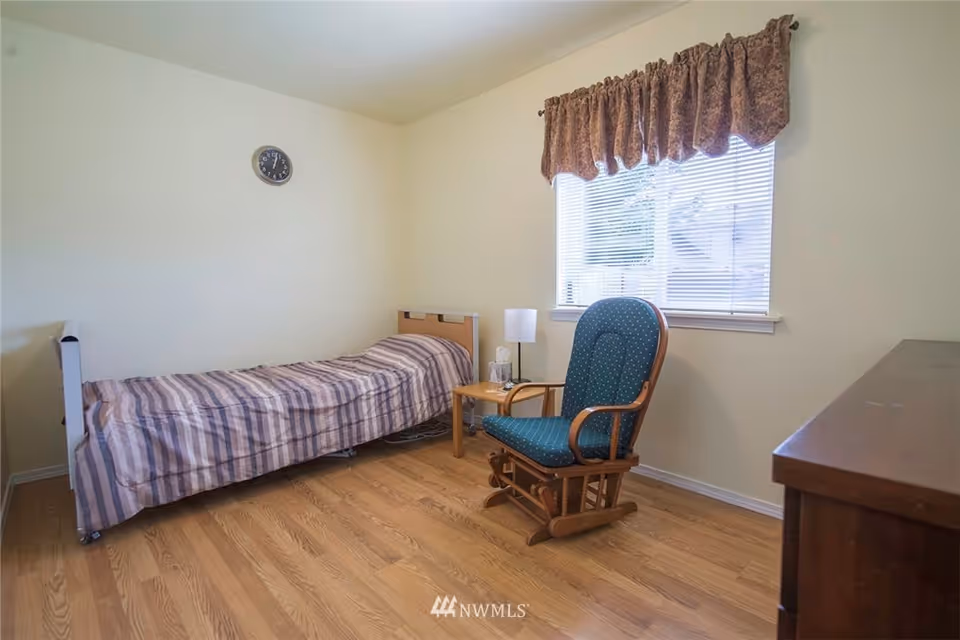 A simple bedroom with a single bed covered in a striped blanket, a wooden rocking chair with green upholstery, a small wooden side table with a lamp and tissue box, a window with blinds and a patterned valance, a wall clock, and wooden flooring.