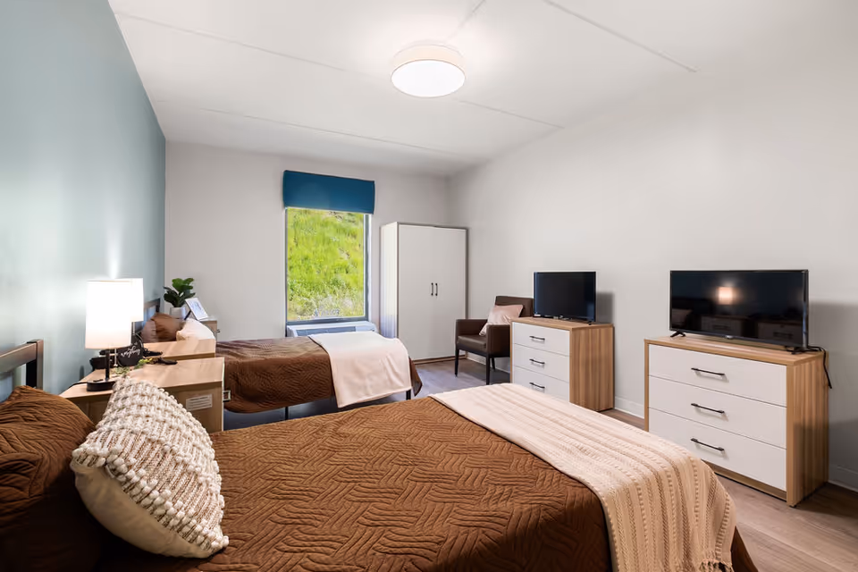 Bright twin bedroom with two beds, dressers, TVs, a chair and a window showing greenery.
