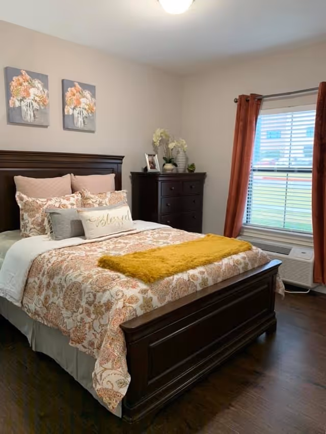 A cozy bedroom with a dark wooden bed frame and matching dresser. The bed is made with patterned bedding in shades of beige and orange, accented with multiple pillows including one with the word 'Blessed'. Above the bed are two floral paintings. A window with red curtains and white blinds lets in natural light, and there is a white air conditioning unit below the window.
